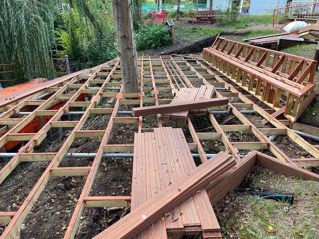 Sub-frame being built around two trees in the centre of the area, and the frame of a long custom bench is to the right of the boardwalk sub-frame.